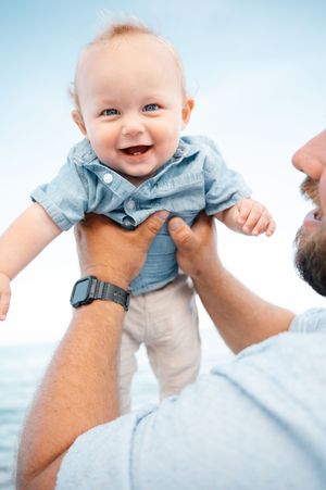 Joyful baby in blue shirt lifted by father against clear sky at beach.