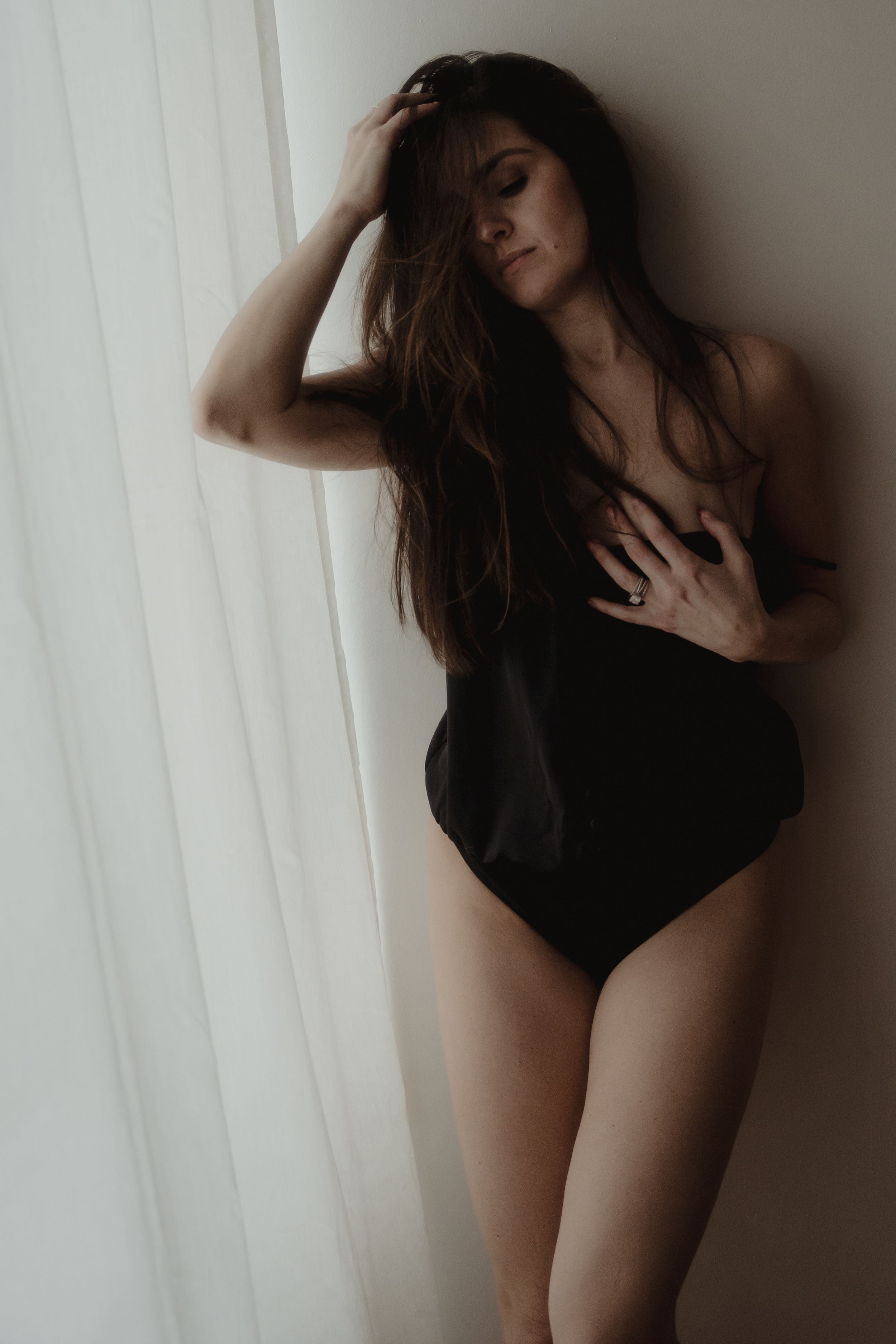 Moody boudoir portrait of woman in black bodysuit against white curtains.