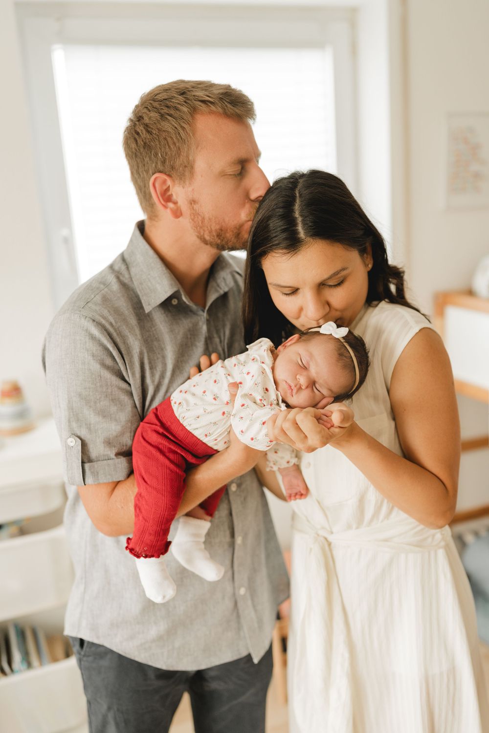 Eltern küssen ihr neugeborenes Baby auf dem Arm - Liebevolle Babyfotografie im eigenen Zuhause in Biberach von Ramona Maier