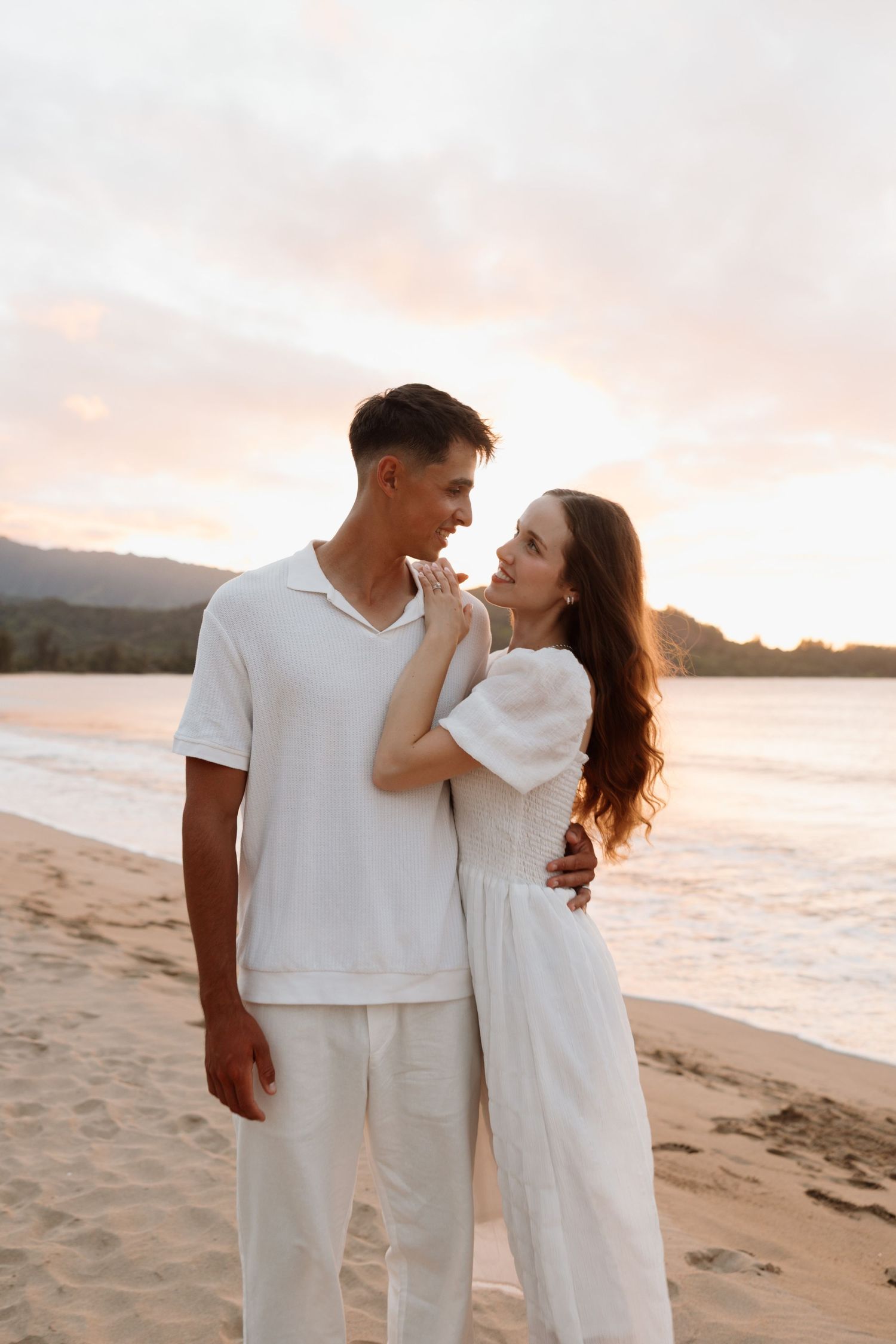 Couple embracing on a beach at sunset with dramatic mountains in the background.