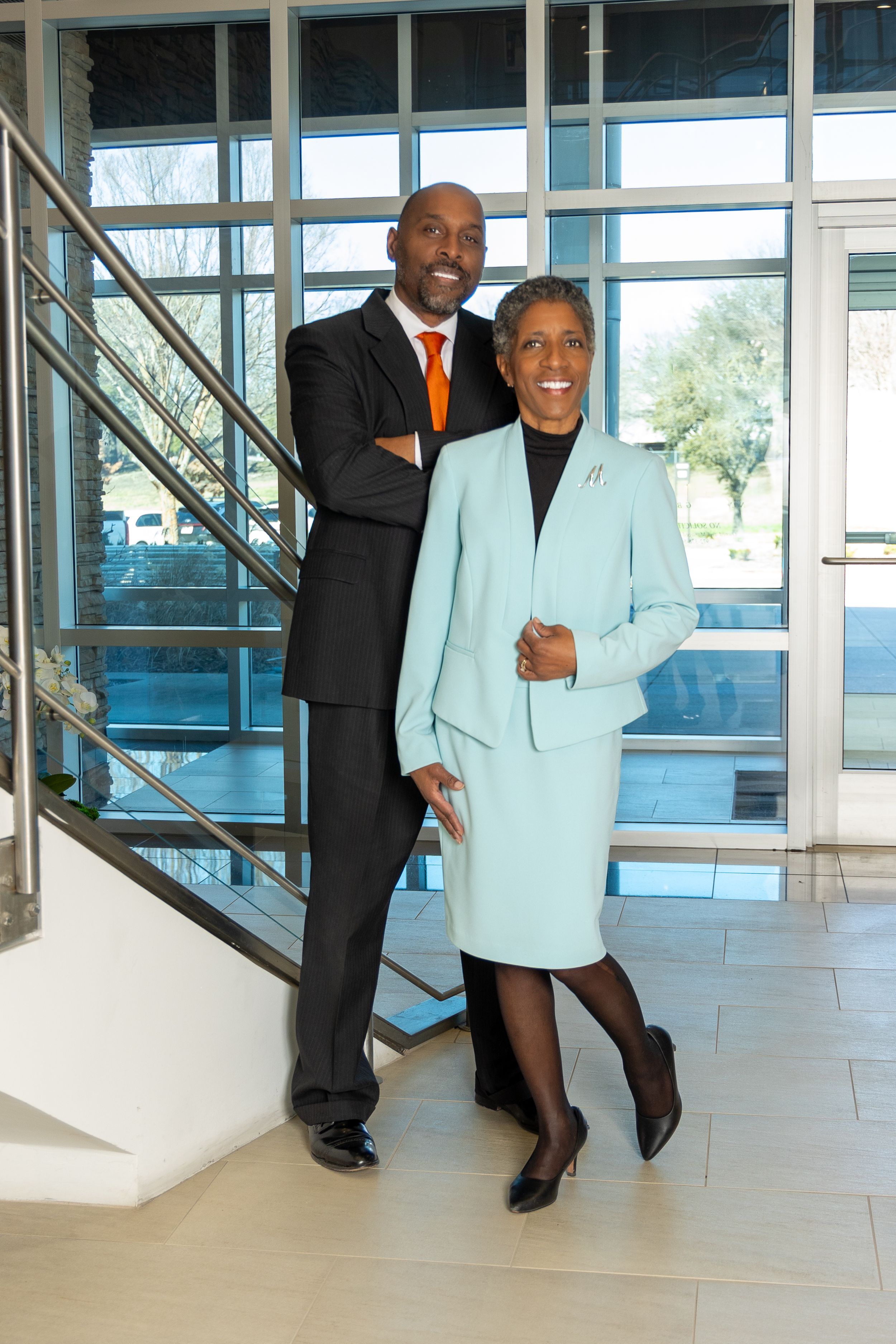 Professional couple in business attire posing by modern glass staircase.