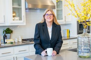 Professional wearing a dark blazer and glasses stands relaxed in a modern white kitchen with yellow floral decor.