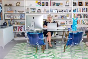 Woman working at glass desk in stylish home office with bookshelves, blue acrylic chairs, and green patterned rug.