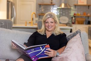 Smiling blonde woman in black dress reading a colorful book on a gray sofa in a modern living room.