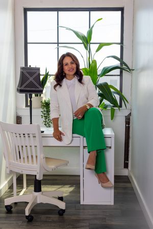 Woman in white blazer and green pants sits on desk in bright office with plants and lamp.