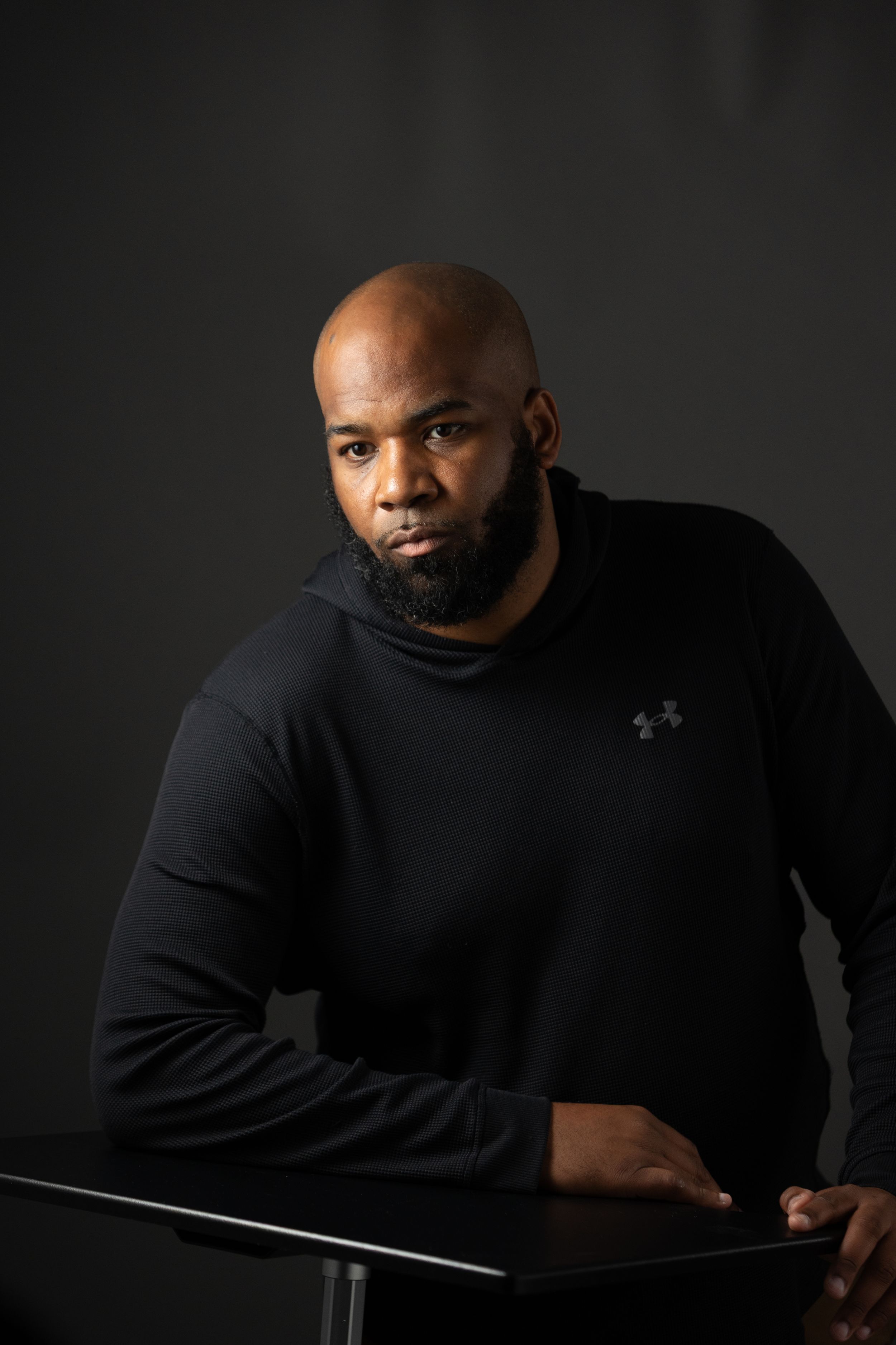 Bald man with beard wearing black Under Armour hoodie poses at table against dark studio background.