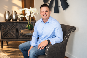 Smiling man in blue shirt sitting in gray chair beside elegant console table with white orchids.