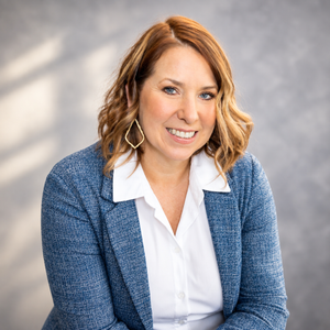 Professional woman in blue blazer and white blouse smiling confidently against a gray studio background.