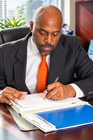 A bald African American businessman in a suit and orange tie signs documents at a desk in a professional office.
