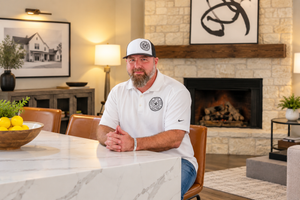 Man in white polo and cap sits at marble kitchen island near stone fireplace in modern home interior.