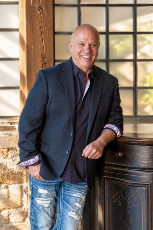Smiling bald man in navy blazer and ripped jeans leaning against ornate post in outdoor setting.
