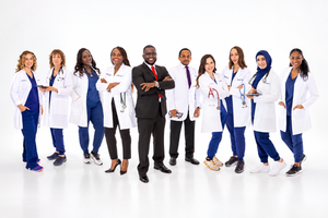 A diverse group of medical professionals in white lab coats pose together smiling, led by a man in a suit.