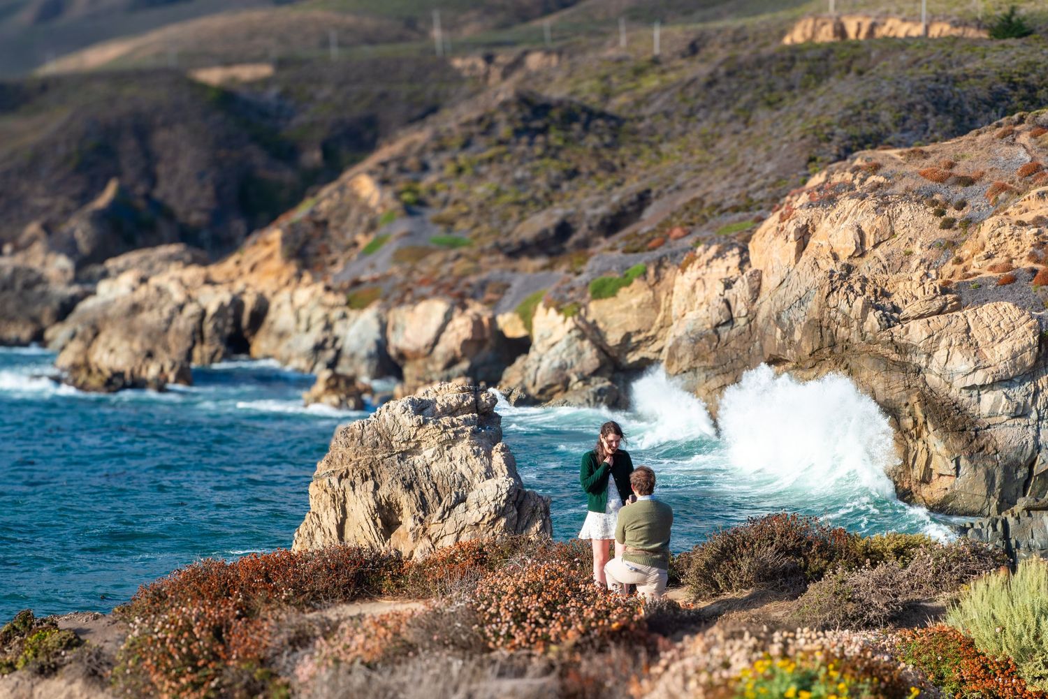 Two people sitting on rocky coastal cliffs watching dramatic ocean waves crash against Big Sur shoreline.