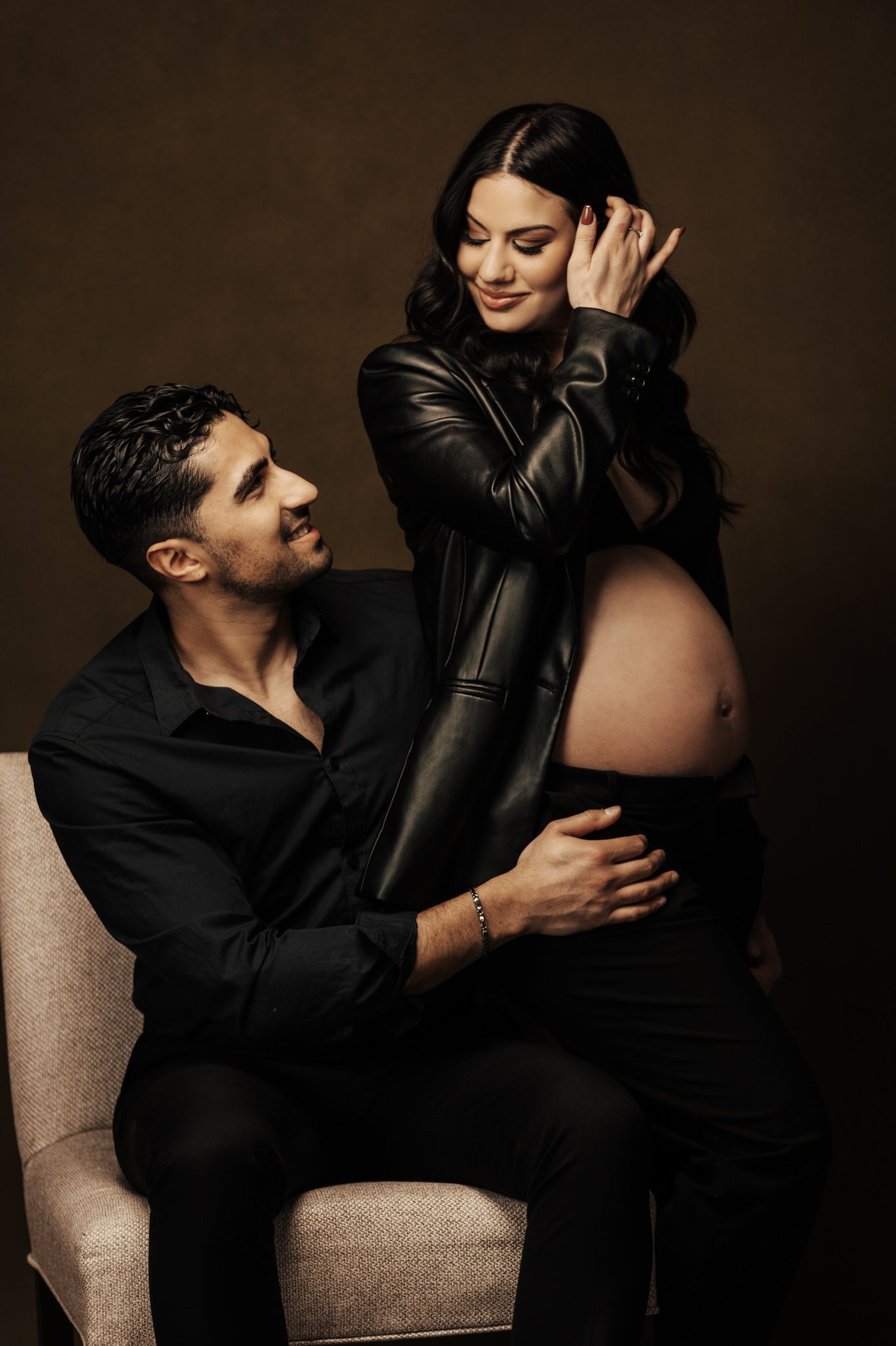 Artistic maternity photography session with couple in black attire on beige furniture.