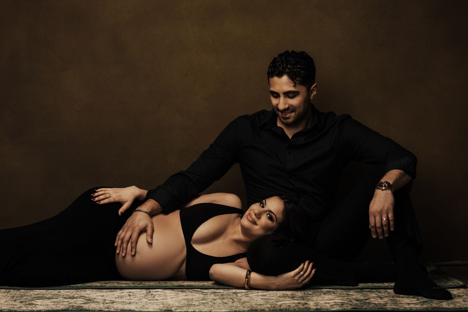 Romantic couple in black clothing poses intimately on dark studio floor.