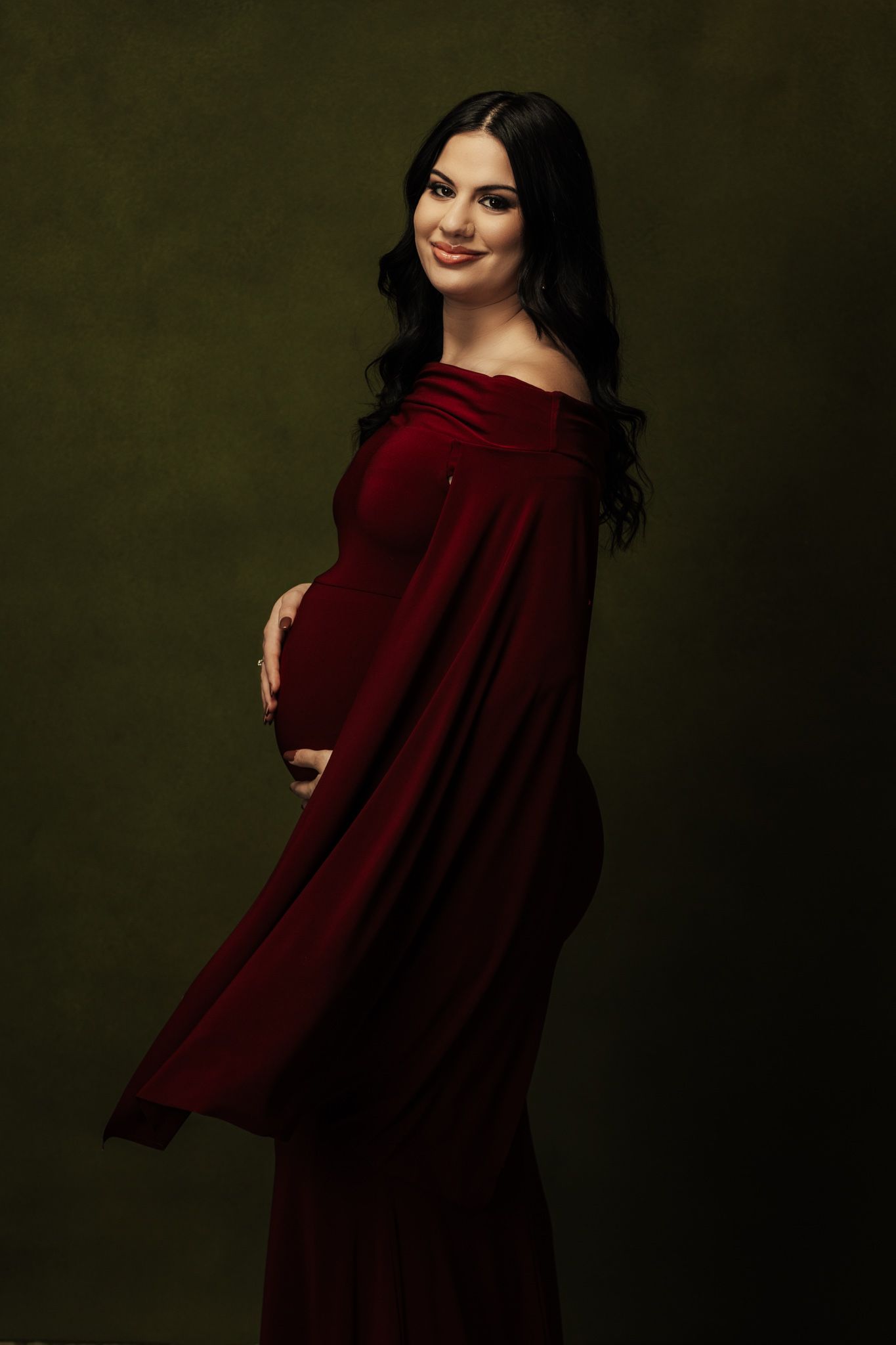 Woman in flowing burgundy dress poses elegantly against dark studio backdrop.