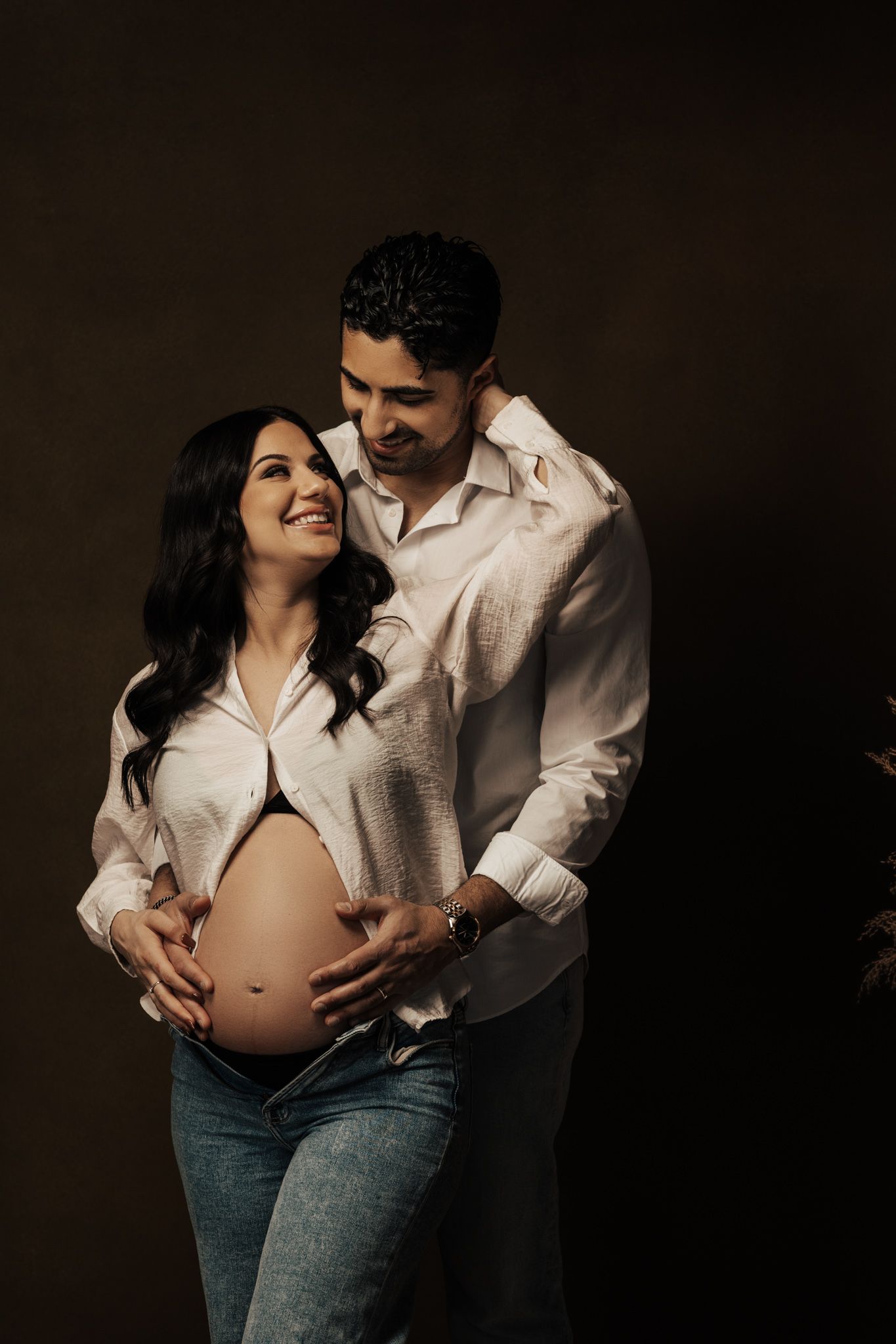 Intimate maternity photo shoot featuring couple in white shirt and jeans against dark background.