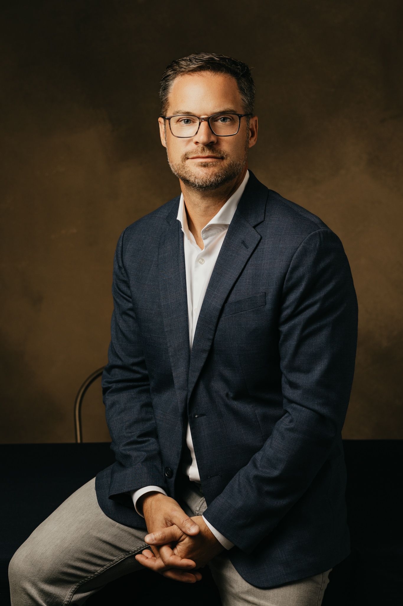 Professional headshot against brown backdrop wearing navy blazer and glasses.