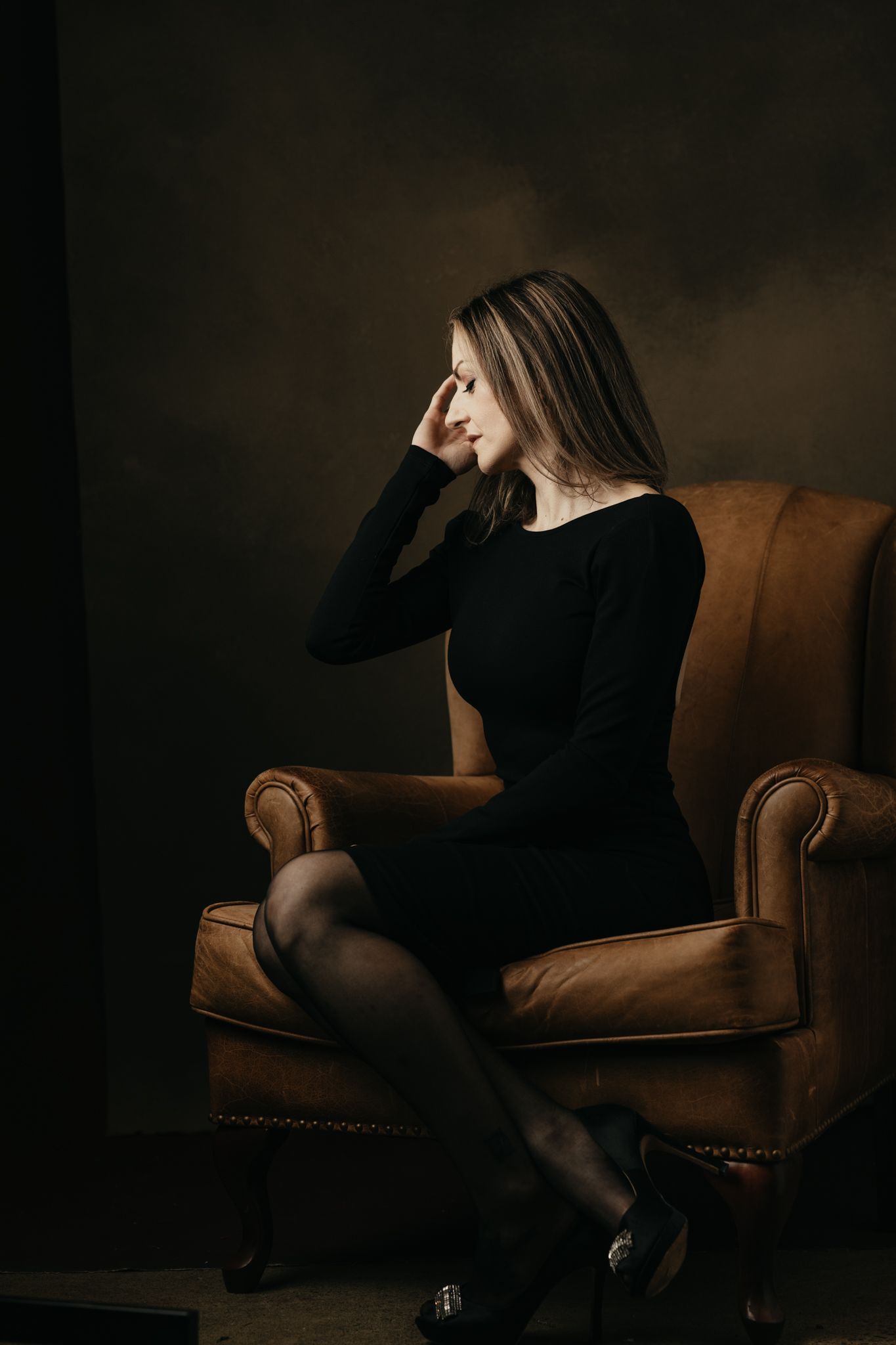 Moody portrait in black dress seated on brown leather armchair with dramatic lighting.