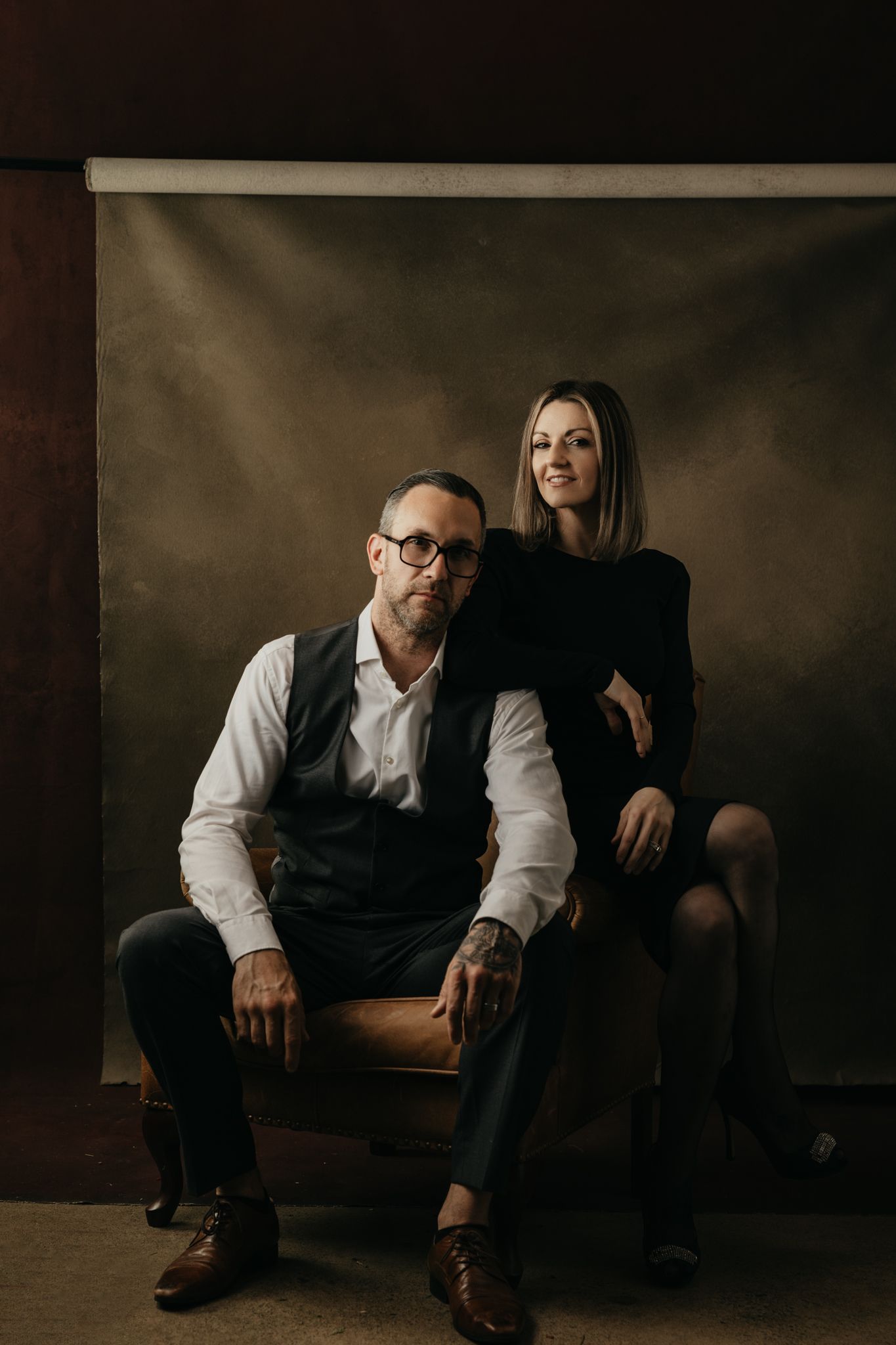 Studio portrait with two people on leather chair against dark backdrop.