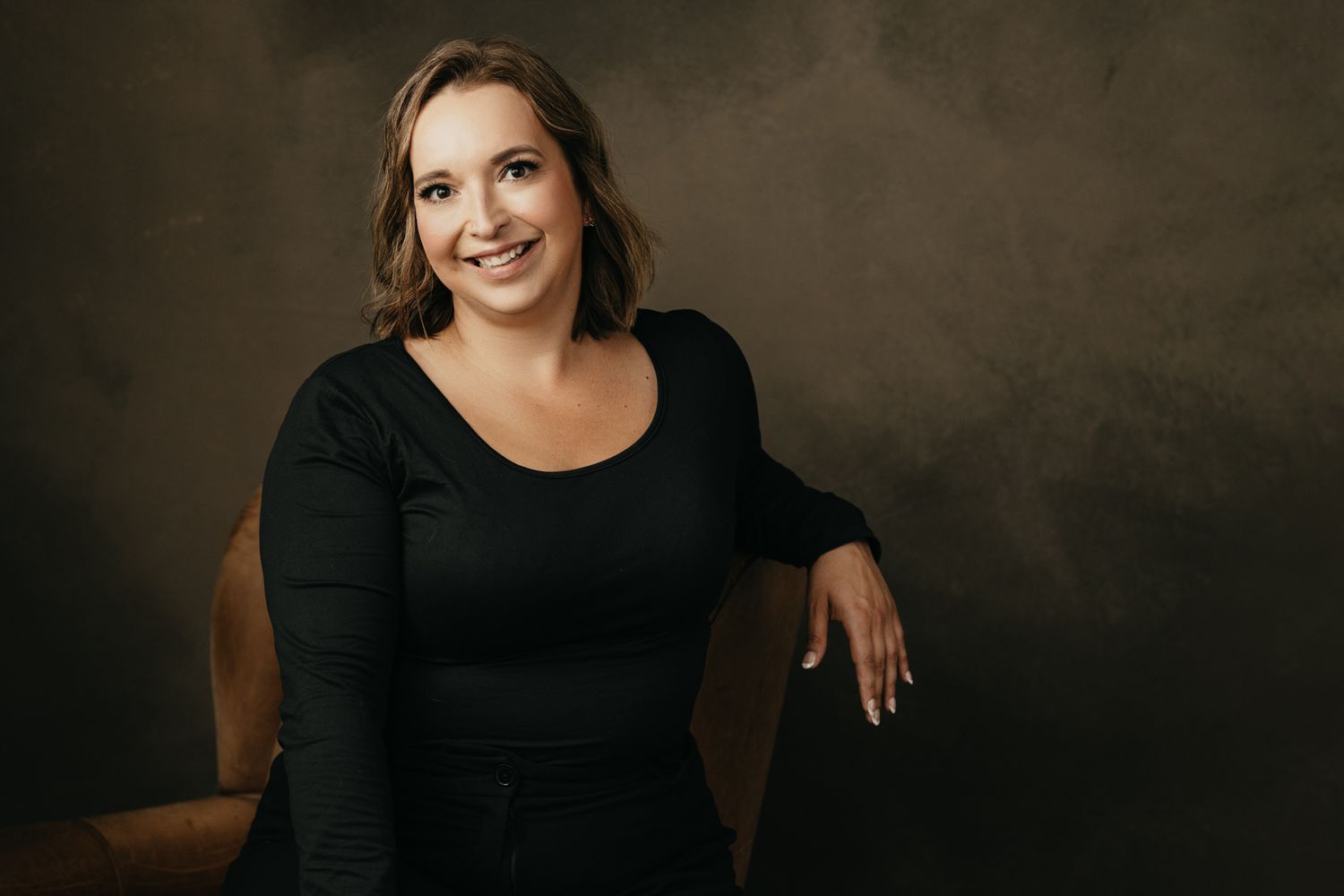 Studio portrait on brown chair with natural lighting and dark background.