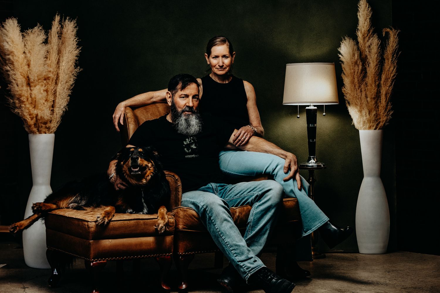A couple relaxes on a leather couch with their dog, flanked by tall white vases filled with pampas grass.