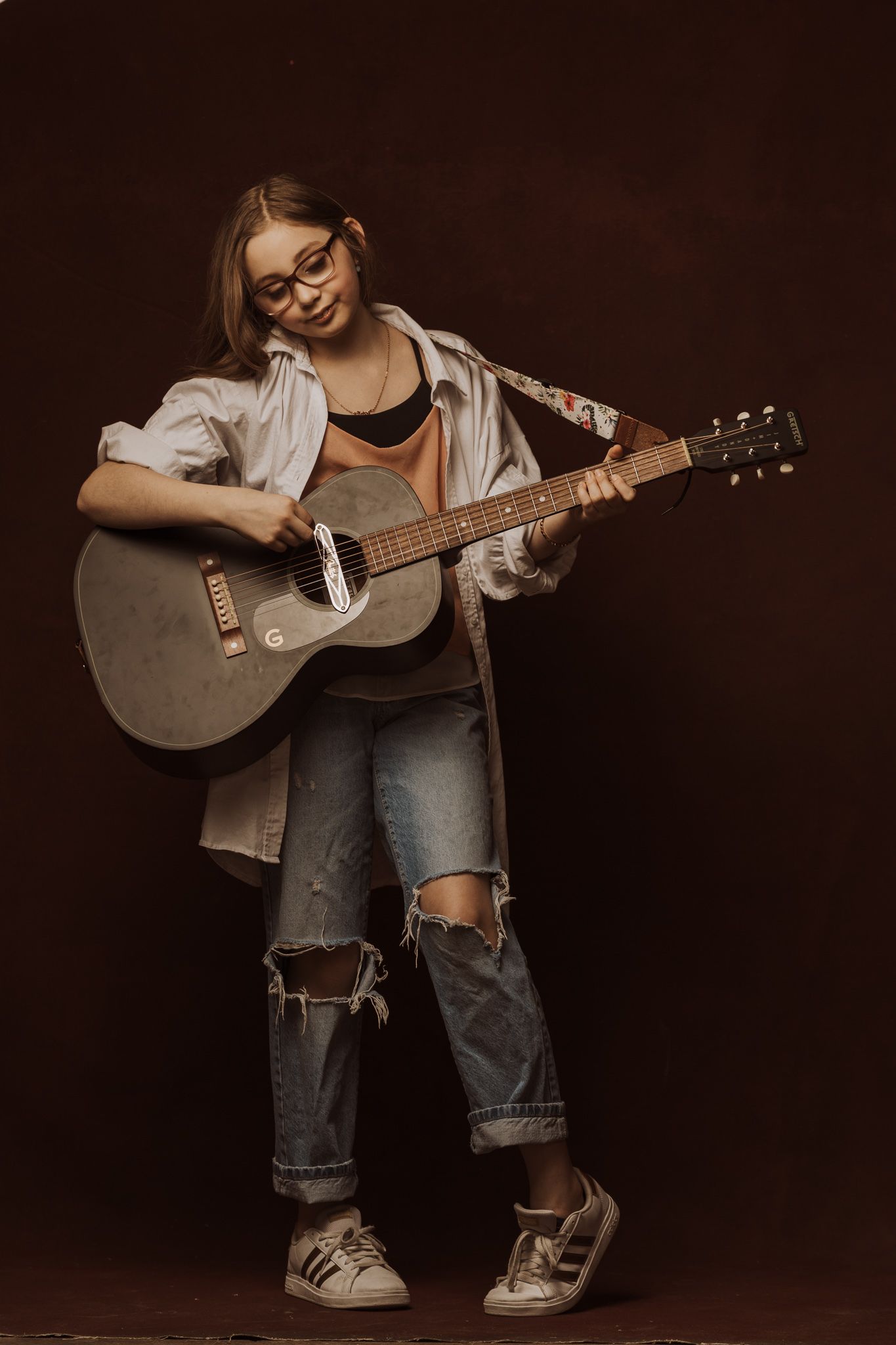 A person in ripped jeans sits in a dark studio playing an acoustic guitar against a moody brown backdrop.