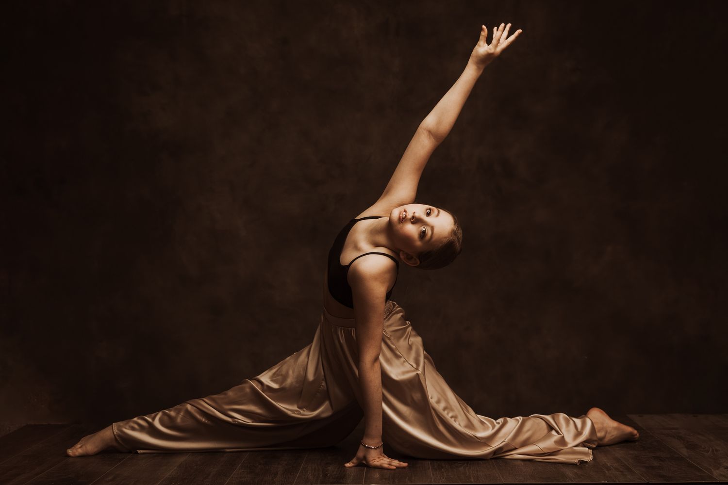 A dancer in a flowing bronze dress performs an elegant backbend pose against a dark background.