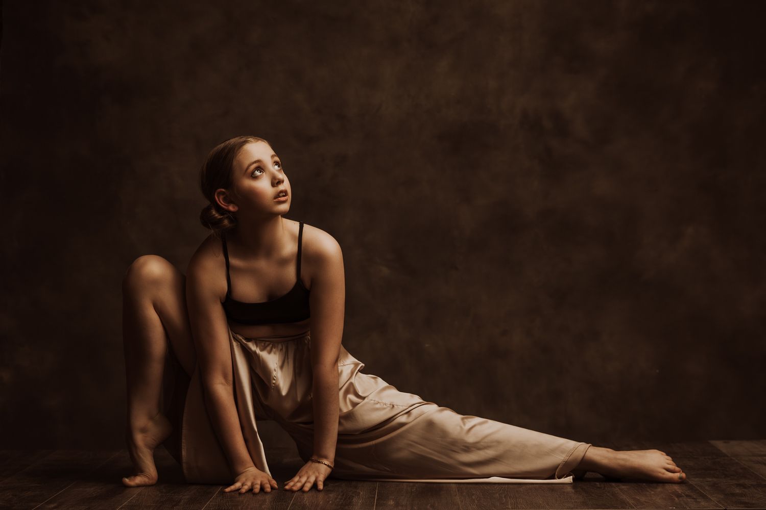 A dancer in a flowing beige dress performs an elegant floor sequence in a dark studio setting.