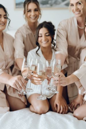 Bridesmaids in blush robes toast with champagne glasses during wedding preparations.