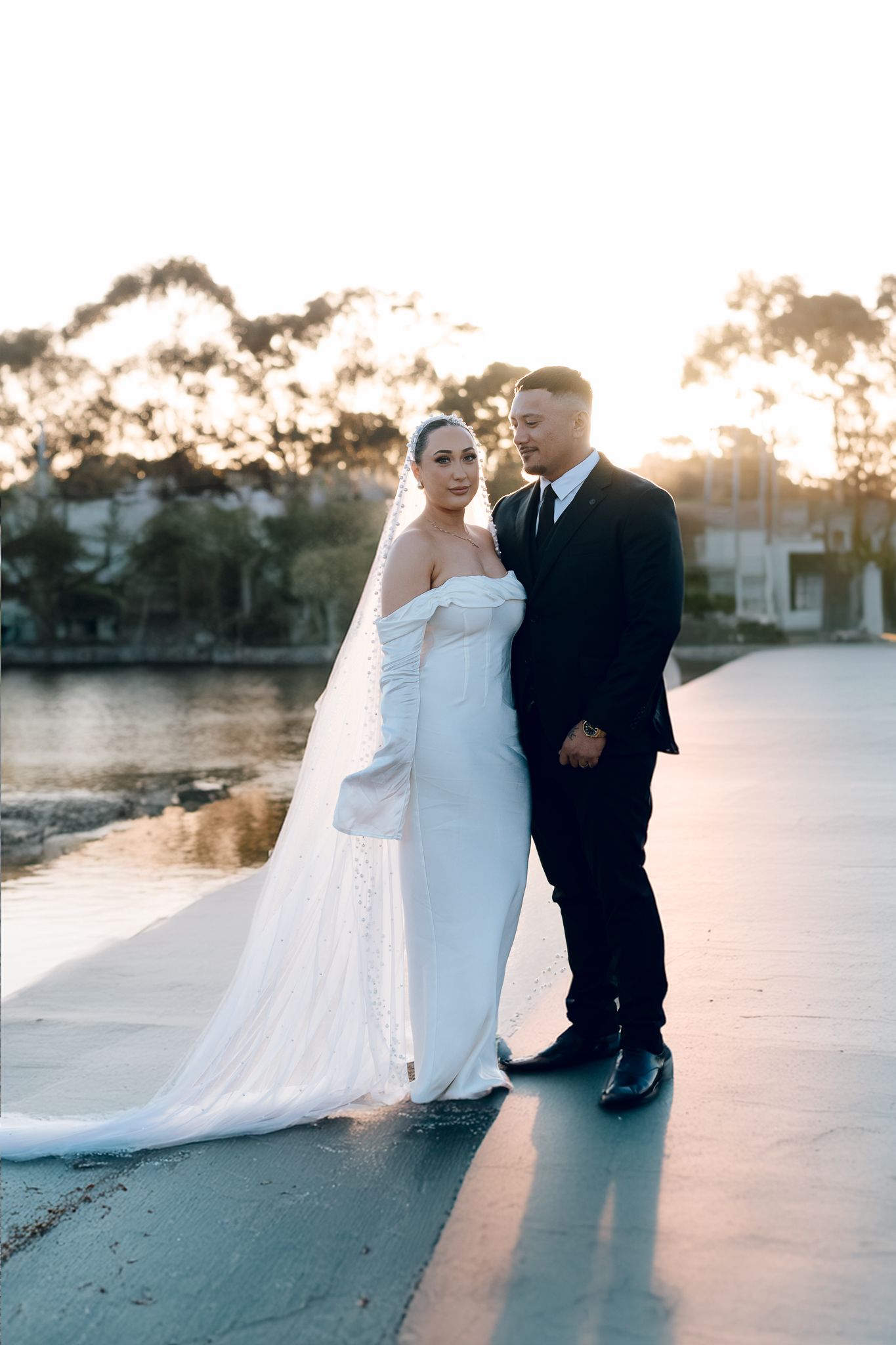 Wedding couple photographed at sunset by a waterfront in formal attire.