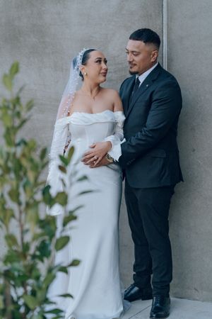 Modern wedding couple posing against concrete wall with greenery.