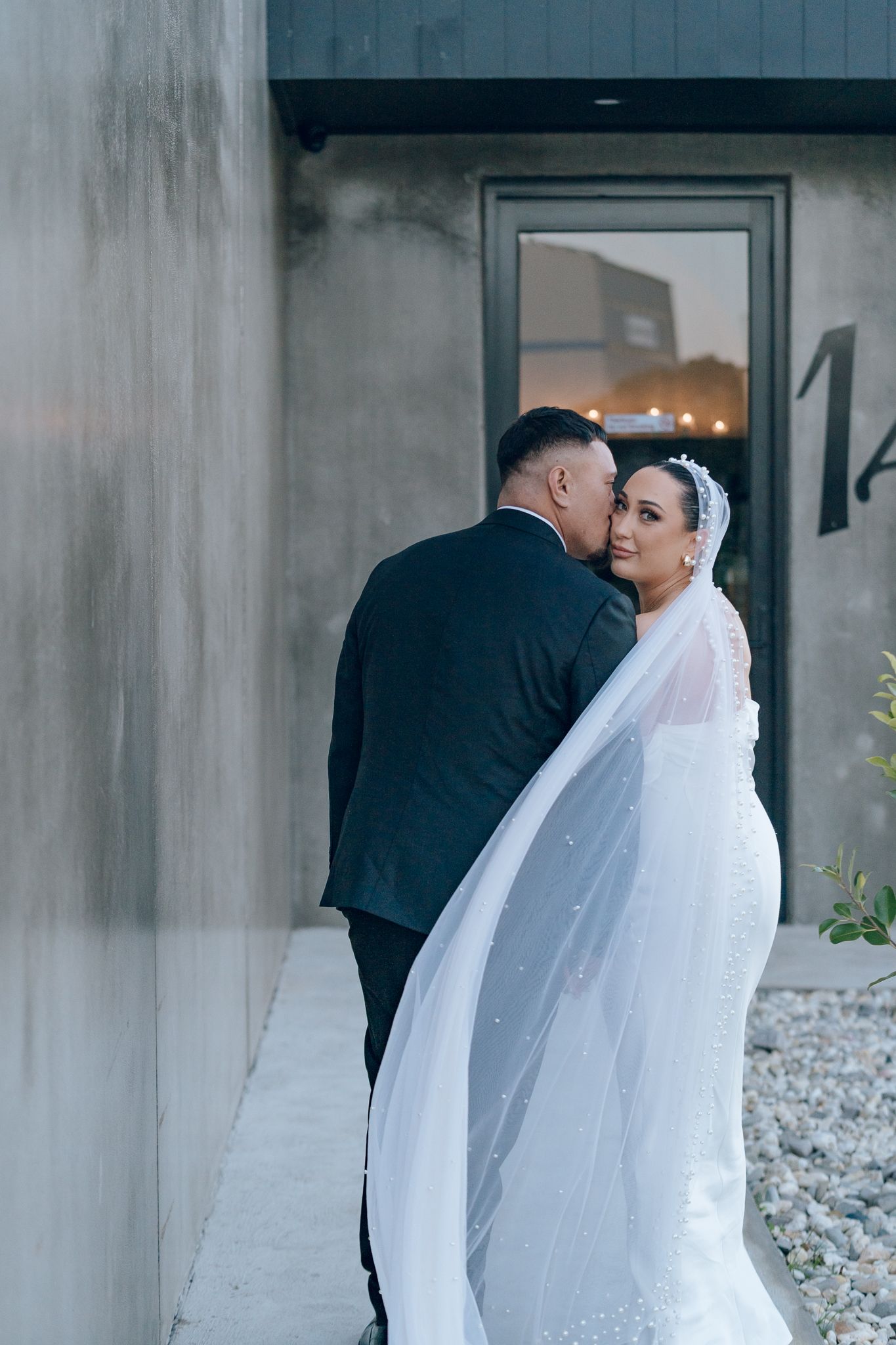 Romantic wedding portrait of couple embracing with flowing white veil against industrial grey wall and modern doorway.