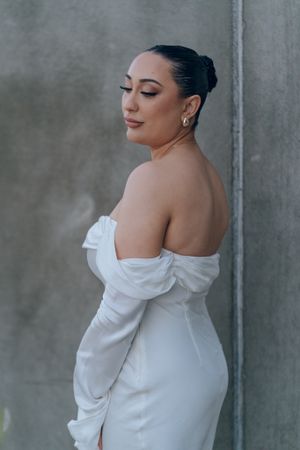 Elegant bride in off-shoulder white wedding dress poses against grey wall backdrop.