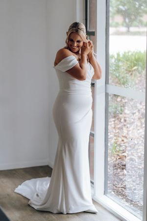 Elegant bride in fitted off-shoulder white wedding gown poses by window.