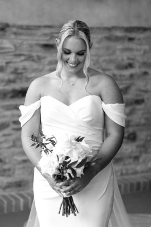 Joyful bride in white off-shoulder dress holding bouquet in black and white portrait.