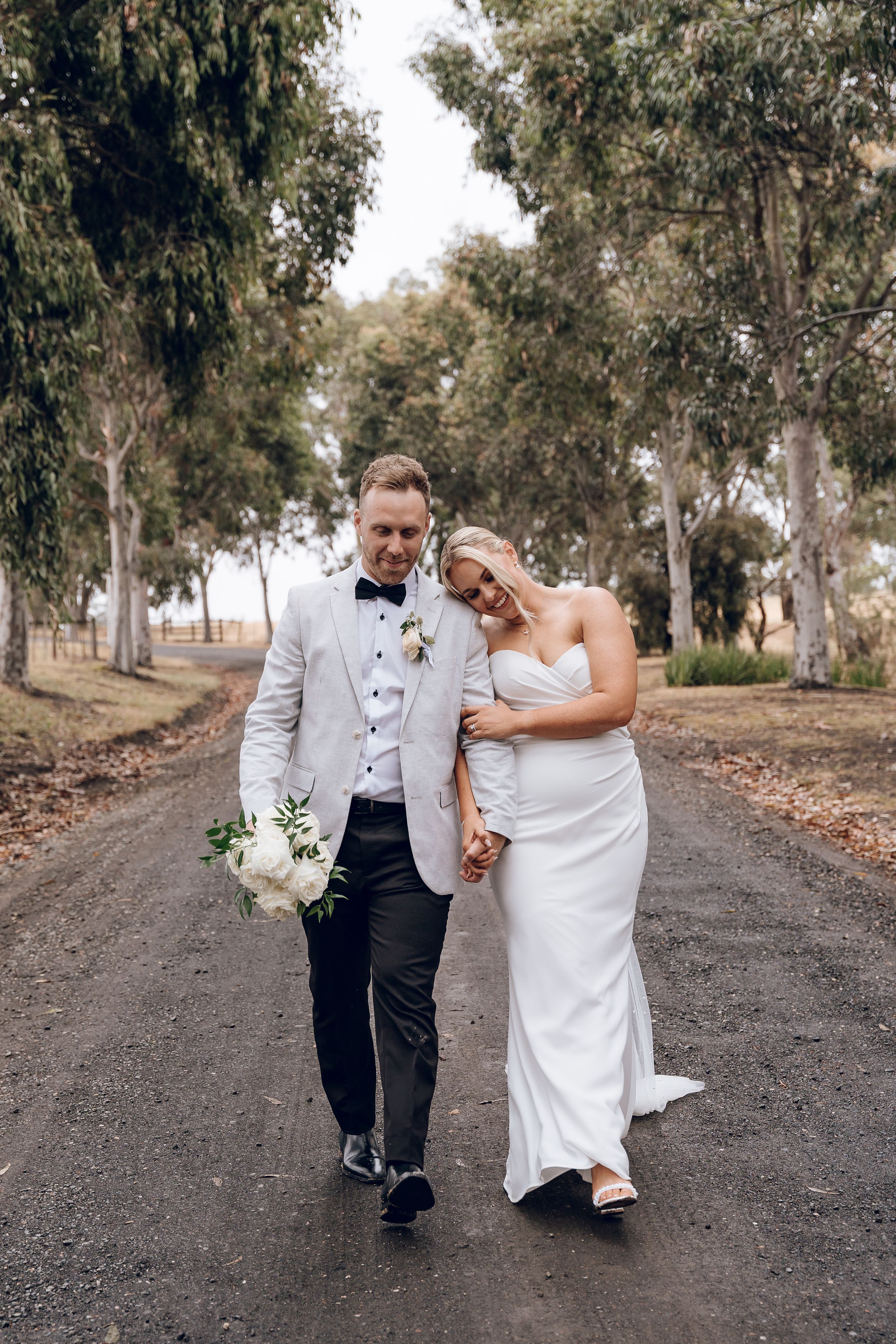 Newlyweds walk together down tree-lined path in elegant wedding attire.