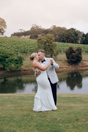 Wedding couple dances by lakeside with vineyard hills in background.