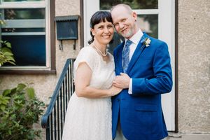A couple in wedding attire embrace and laugh together in front of a light colored building exterior.