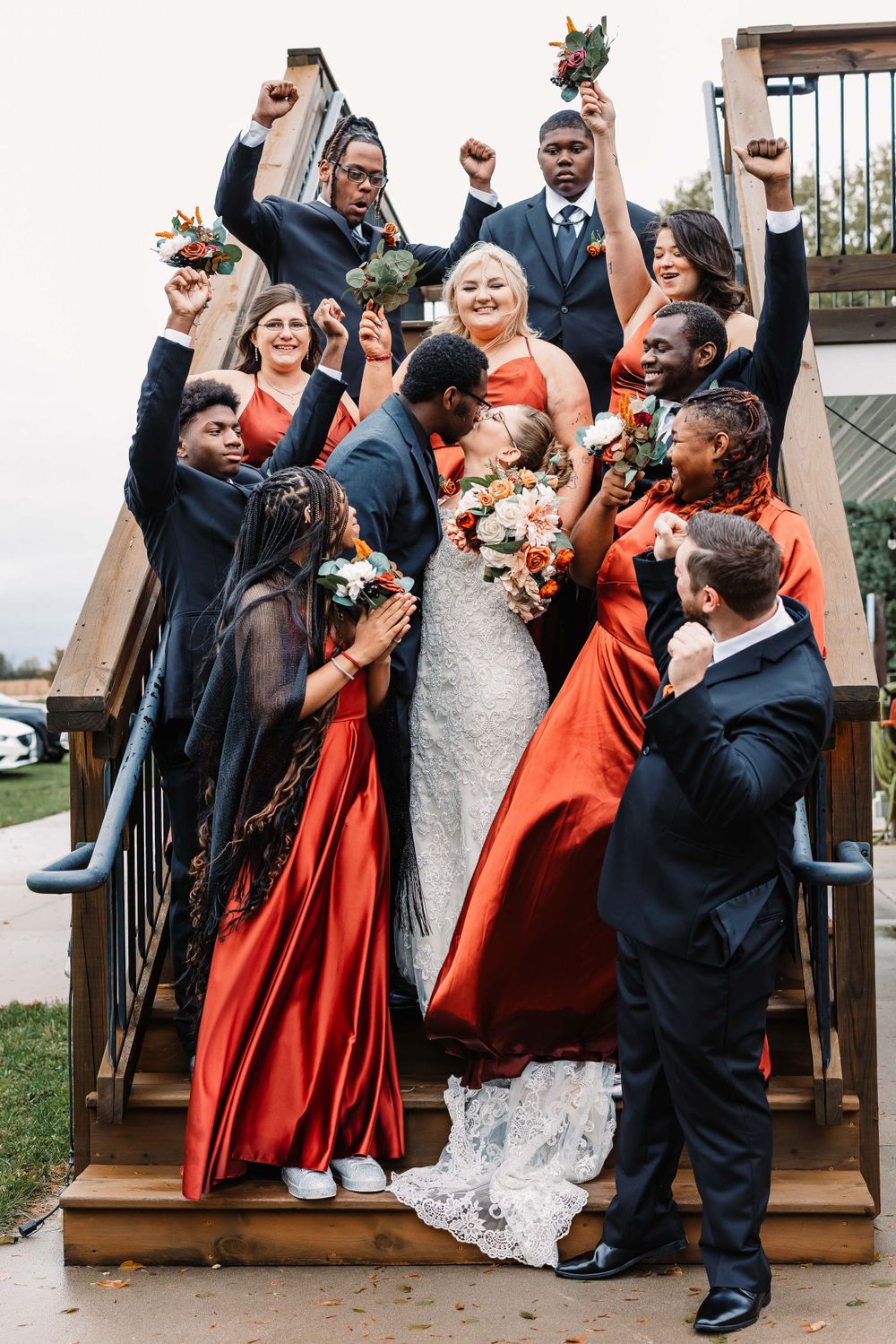 Wedding party poses joyfully on wooden stairs with bridesmaids in red dresses and groomsmen in dark suits.