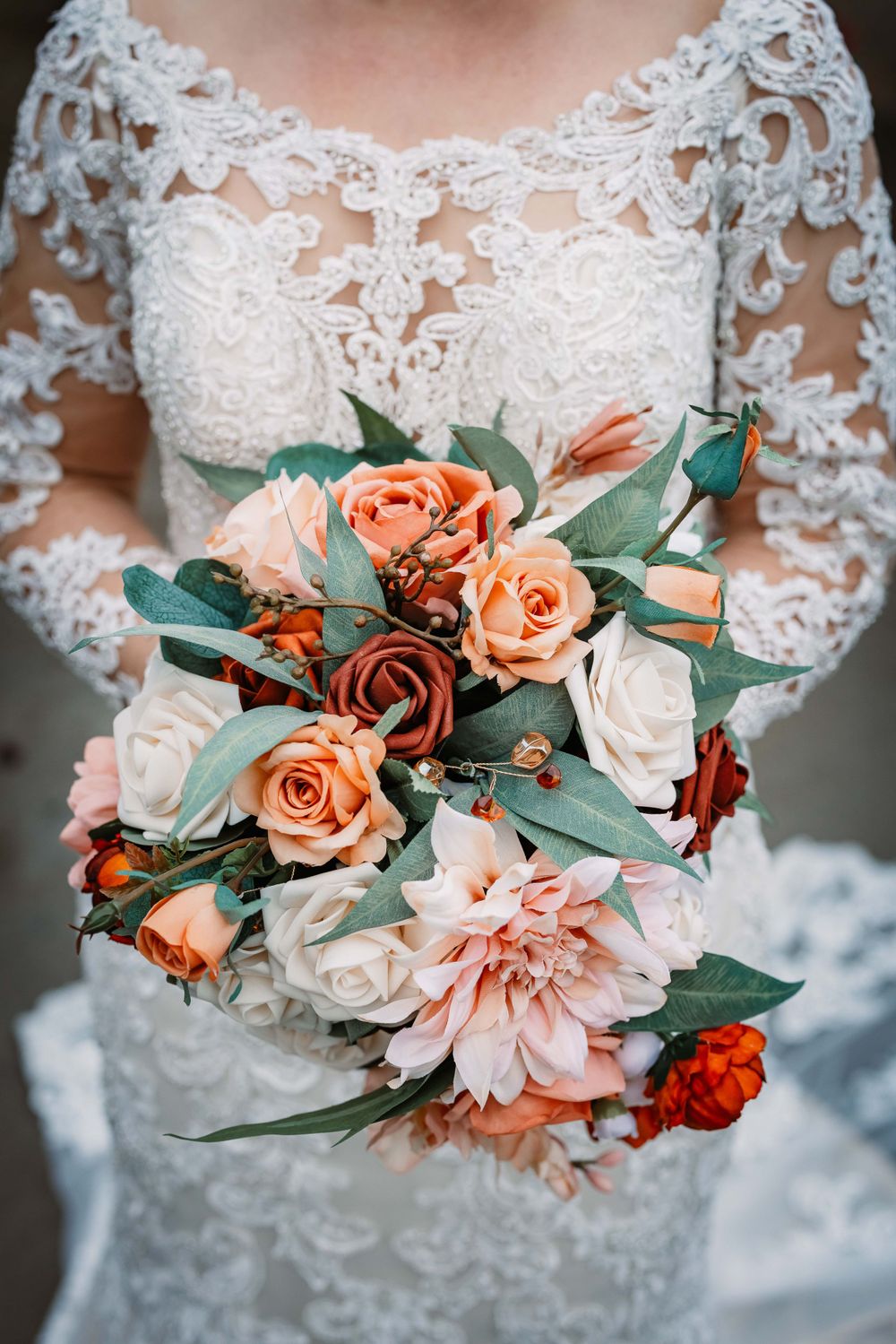 Close-up of bridal bouquet featuring peach roses, white flowers and eucalyptus against lace wedding dress.