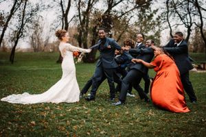 Wedding party plays tug-of-war on grassy lawn with bride in flowing white dress.