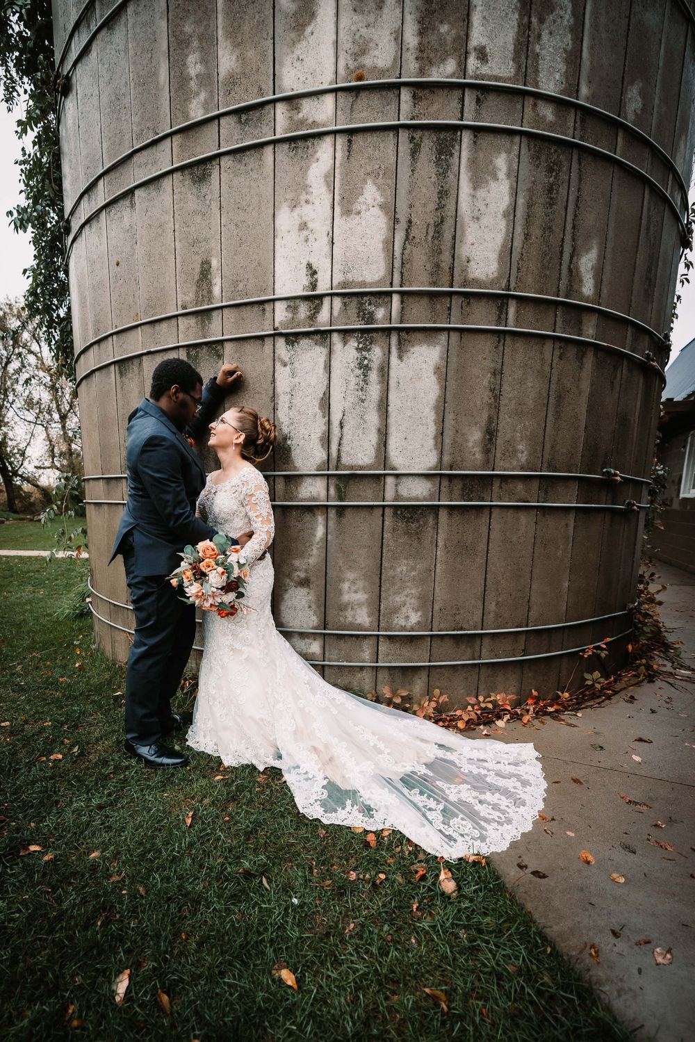 Wedding couple poses against large rustic silo structure with trailing lace dress and fall bouquet.