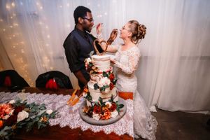 Newlyweds share a playful moment while feeding each other wedding cake beside a decorated dessert table.