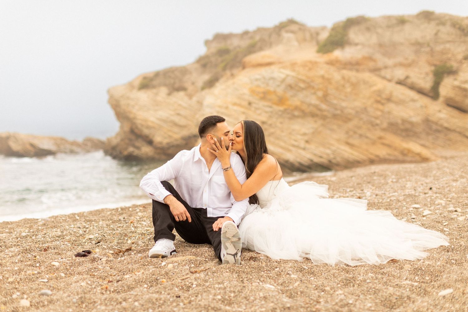 A romantic coastal wedding photo with waves crashing against rocky cliffs in the background.