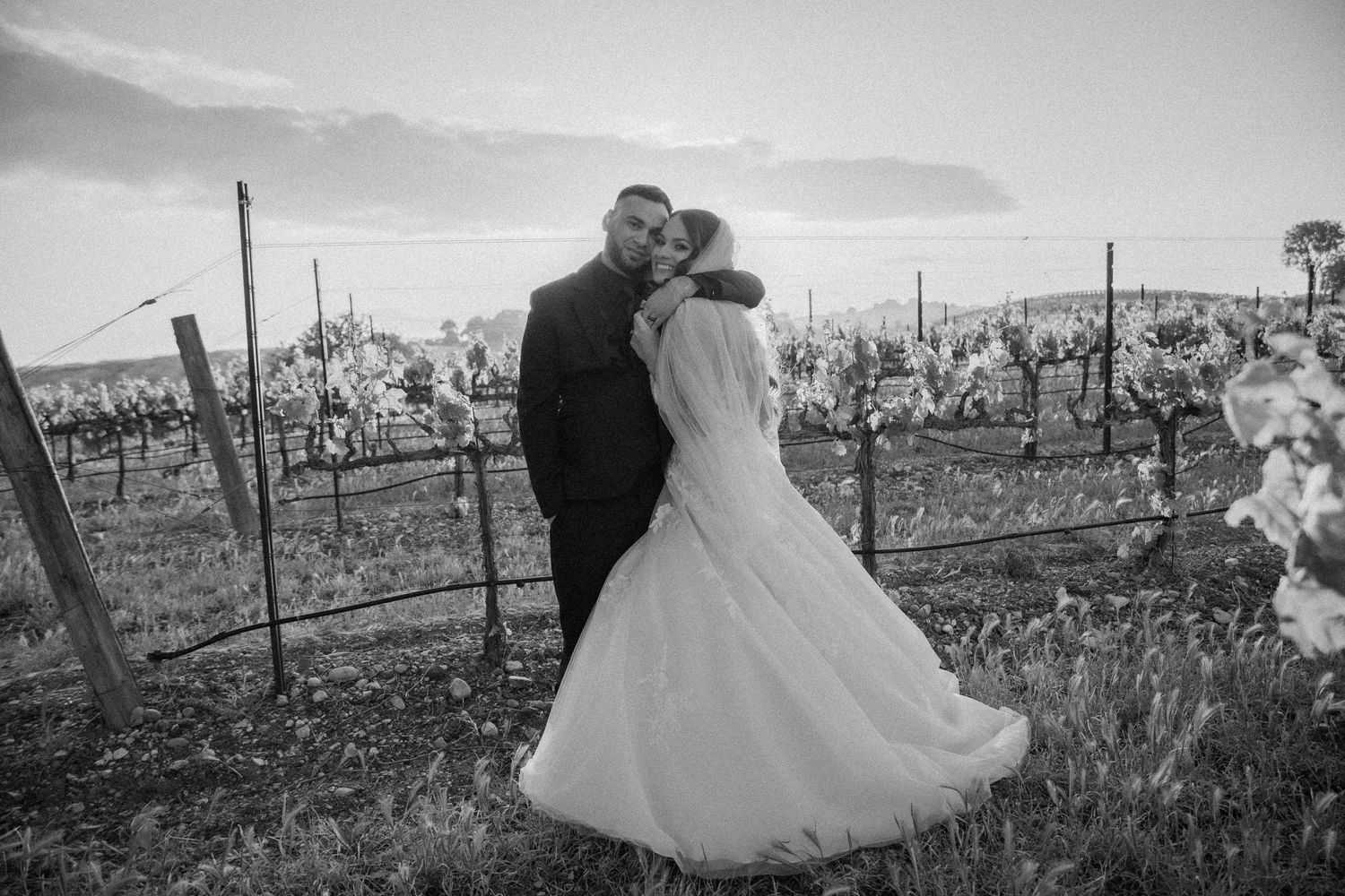 A black and white wedding photo with couple embracing in a vineyard landscape during winter.