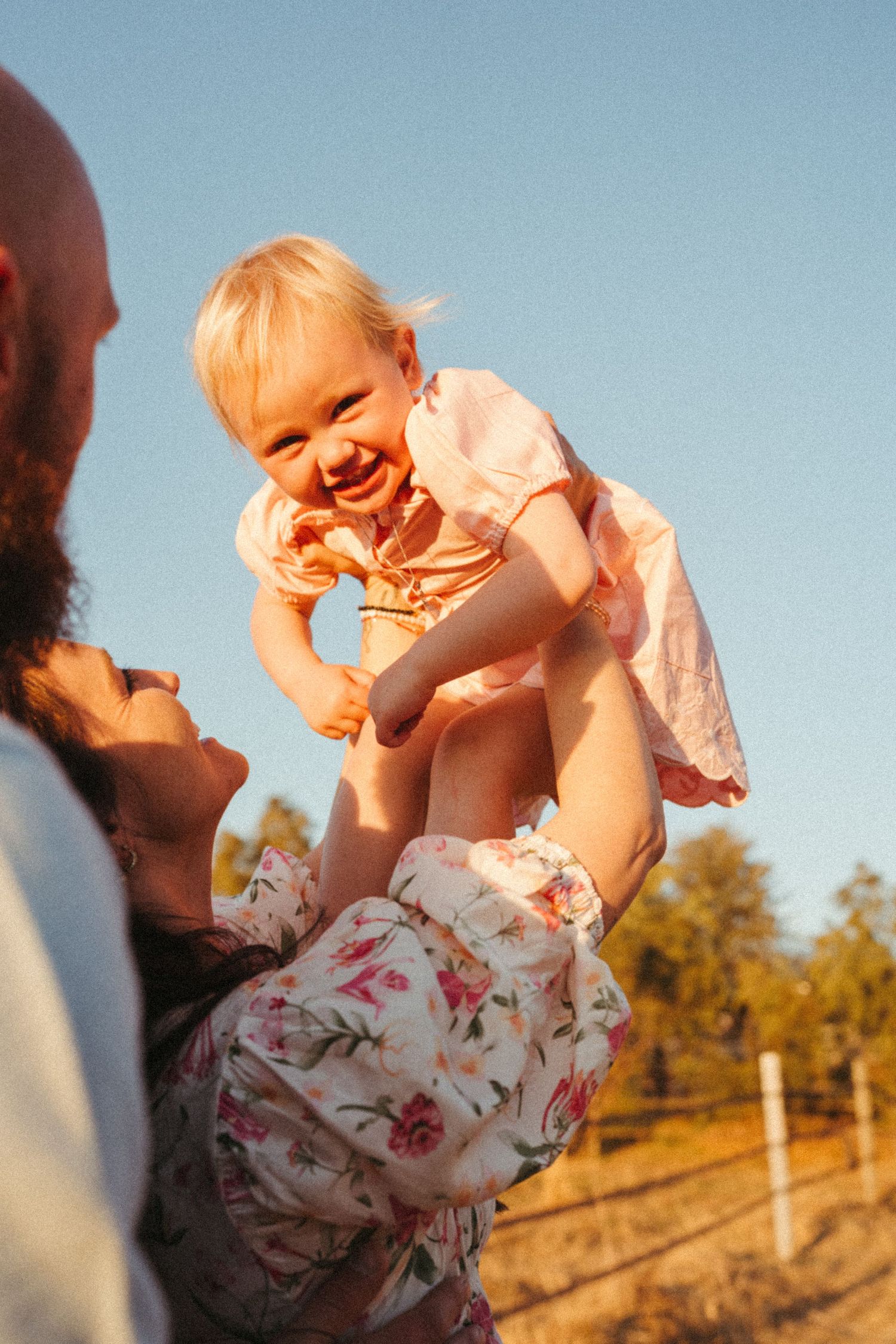 A laughing baby being lifted up by parents during a sunset photoshoot.