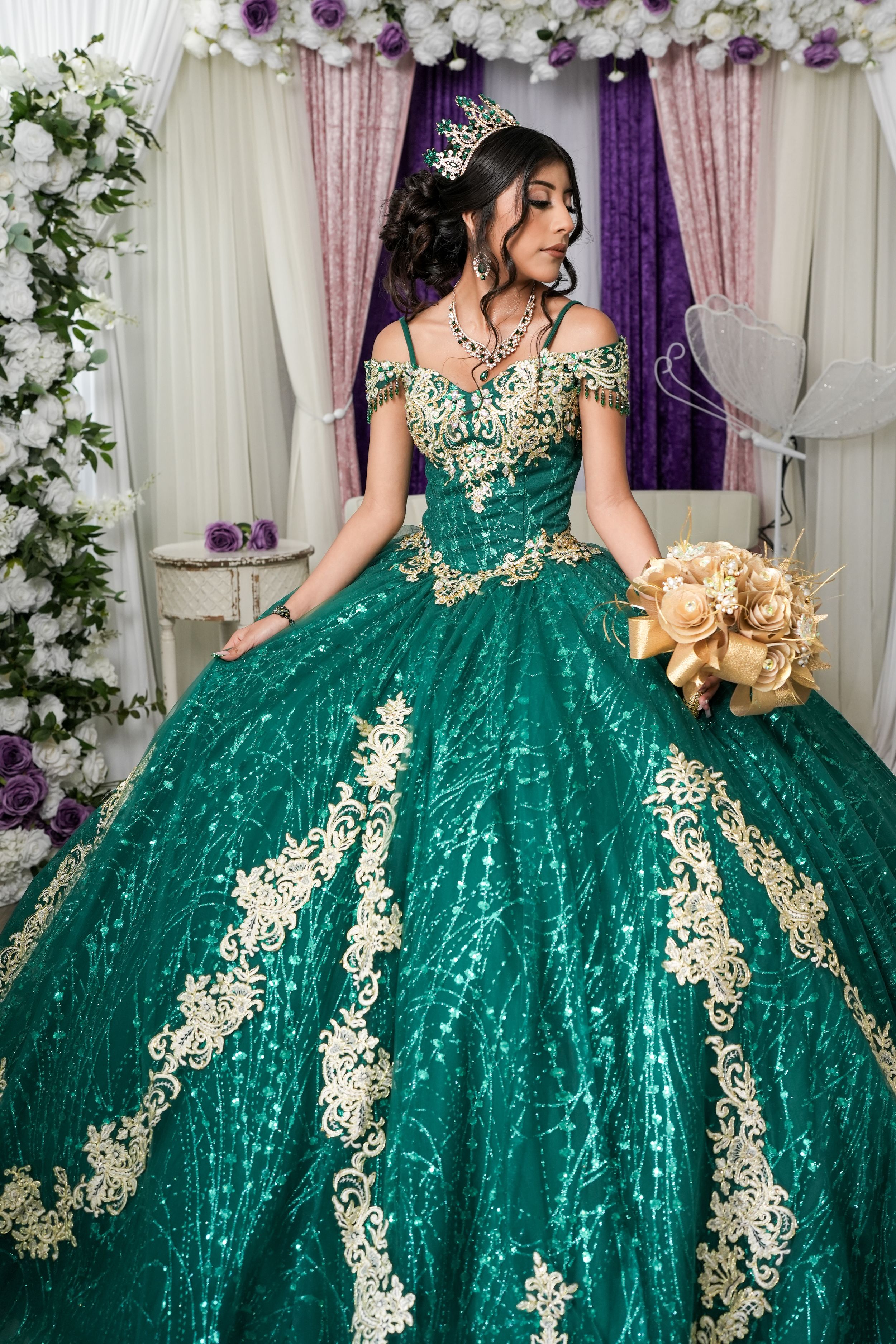 Emerald green quinceañera ball gown with gold lace appliques and off-shoulder design against purple and white backdrop.