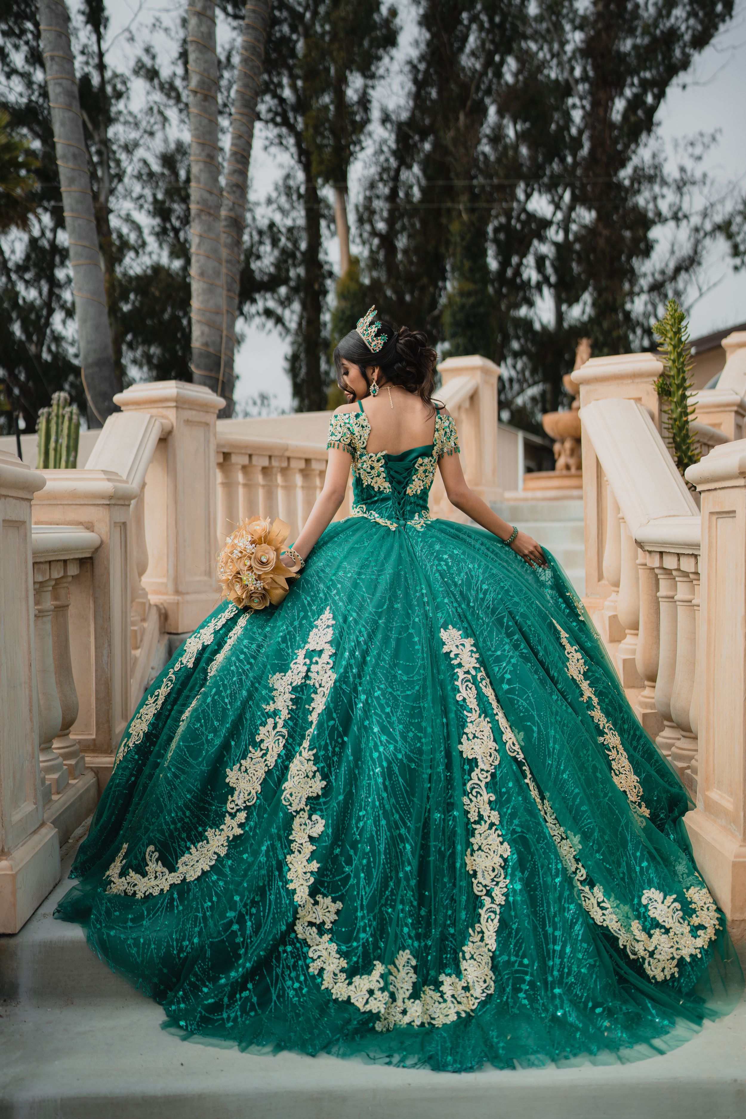 A stunning emerald green quinceañera ball gown with intricate gold embroidery details spreads across the floor in a majestic display.