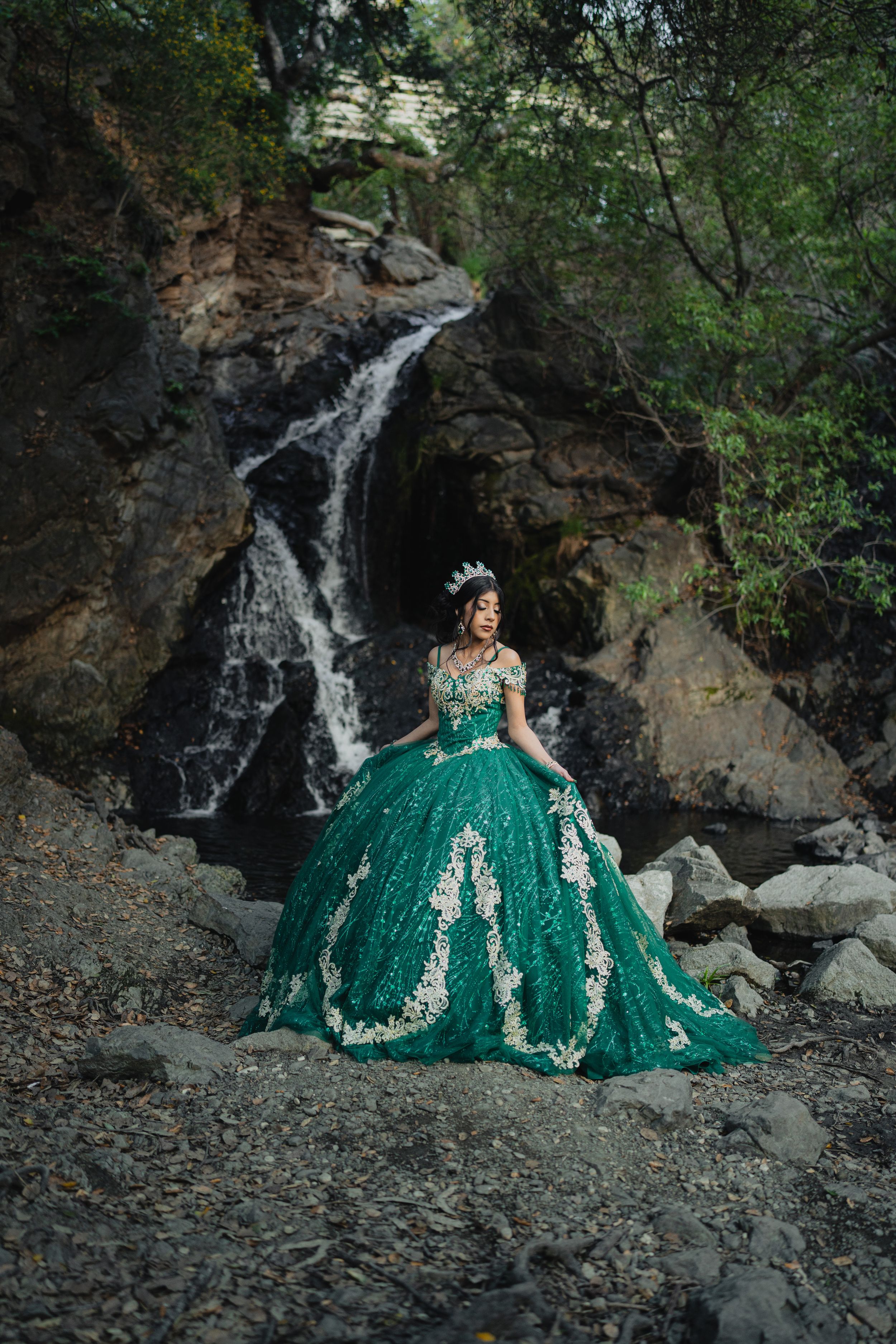 Woman in emerald green quinceañera dress poses elegantly near a cascading waterfall in a natural forest setting.